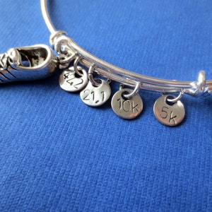 May include: Silver-toned expandable bangle bracelet with a running shoe charm and four round charms. The round charms are engraved with the numbers and distances: 42.2, 21.1, 10k, and 5k. The bracelet is on a blue background.