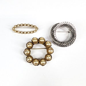 May include: Three brooches, one with a circle design, one with a row of pearls, and one with a circle of pearls.
