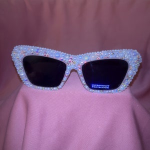 May include: White cat-eye sunglasses embellished with pearls and iridescent beads. The frames have dark lenses and the text "ULTRAVIOLET PROTECTION" is visible on the lens. The sunglasses are set against a pink background.