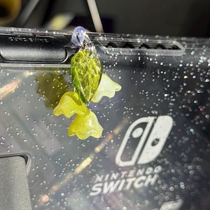 May include: A green and yellow glass charm with a leaf and flower design hangs from a Nintendo Switch console case. The case is clear and has a glittery finish. The text "Nintendo Switch" is visible on the case.