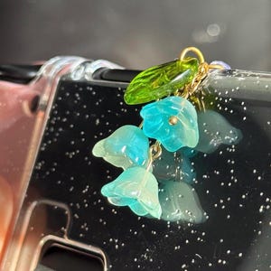 May include: A delicate phone charm featuring a cluster of translucent bluebell flowers and a green leaf. The flowers are suspended from a gold-colored chain, adding a touch of elegance to the accessory.