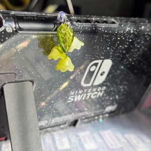 May include: A green and yellow leaf charm with a silver loop, hanging from a clear case for a Nintendo Switch.