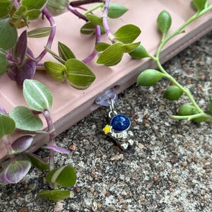 May include: A silver astronaut charm with a blue and sparkly space helmet and a yellow star. The charm is attached to a pink book with green plants.