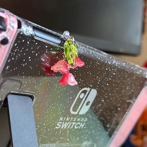 May include: A pink Nintendo Switch console with a clear case and a floral charm attached to the top. The charm features pink and green flowers.
