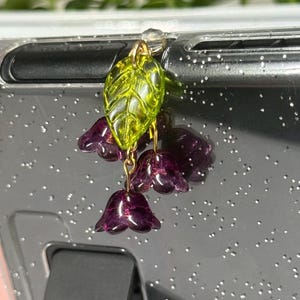 May include: A green leaf charm with purple flower charms hanging from it. The charms are attached to a clear phone case.