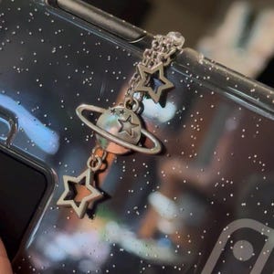 May include: A silver charm with a planet and stars hanging from a chain. The charm is attached to a clear phone case with glitter. The case has the Nintendo Switch logo on it.