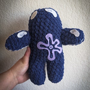May include: A blue crocheted plush toy with a purple flower design on the front. The toy has white felt accents on the head and arms.