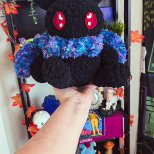 May include: A black crocheted plush toy with large red eyes and a blue and purple fuzzy scarf. The toy is being held in a person's hand.