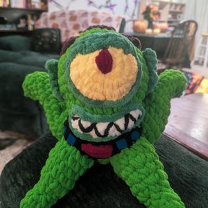 Puede incluir: Un monstruo verde de ganchillo hecho a mano con un ojo grande, iris amarillo y pupila granate. Tiene boca negra y blanca, detalles azules y brazos extendidos. El peluche es una creación caprichosa y única.