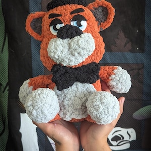 Op de afbeelding: Een gehaakt pluche speelgoed van Freddy Fazbear uit de Five Nights at Freddy's videogame-serie. Het speelgoed is bruin en oranje met een zwarte hoed en strik. Het speelgoed heeft een vriendelijke uitdrukking met blauwe ogen en een zwarte neus.
