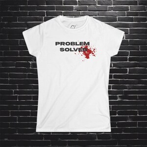 Op de afbeelding: Wit T-shirt met de tekst "PROBLEM SOLVER" in donkergrijze letters en een rood spetterontwerp. Het shirt heeft een ronde hals en korte mouwen. De achtergrond is een donkere bakstenen muur.