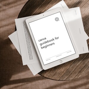 Könnte beinhalten: Ein weißes Tablet-Computer mit einem schwarzen Bildschirm, auf dem ein Canva-Handbuch für Anfänger angezeigt wird. Der Text "canva guidebook for beginners" wird auf dem Bildschirm angezeigt. Das Tablet liegt auf einem Holztisch mit weißem Papier darunter.