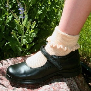 May include: A black leather Mary Jane shoe with a strap and buckle closure. The shoe is paired with a light beige ruffled sock. The background features green foliage and a stone.