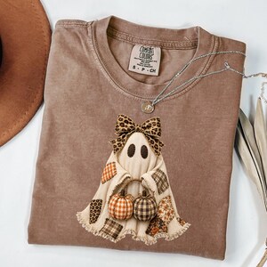 May include: A brown short-sleeve t-shirt with a Comfort Colors label at the neck. The shirt features a graphic of a ghost wearing a leopard print bow, holding two small pumpkins, and decorated with various patterned fabric patches at the hem.