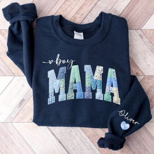 May include: Navy blue sweatshirt with "boy MAMA" in white script and patchwork letters. The sleeve reads "Oliver" with a heart. The patchwork letters are various shades of blue and green.