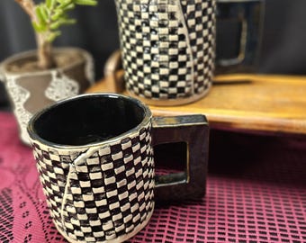 Puede incluir: Dos tazas de cerámica con un patrón de cuadros blancos y negros. Las tazas tienen un asa cuadrada y un interior negro. Las tazas están sobre un mantel de encaje rosa con una planta en el fondo.