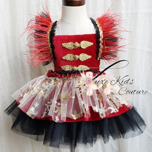 May include: Red velvet dress with gold buttons and black and white tulle skirt with gold stars. The dress has red feather trim on the shoulders.  The dress is on a white mannequin.  The text "Luxe Kids Couture" is visible in the image.