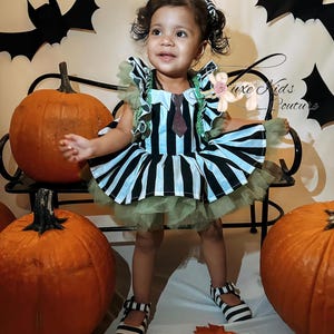 Peut inclure: Une petite fille porte une robe rayée noire et blanche avec une cravate scintillante et des détails verts. La robe a une jupe en tulle et est assortie de chaussures rayées noires et blanches. Le fond comprend des citrouilles, des chauves-souris et le texte "Luxe Kids Couture".