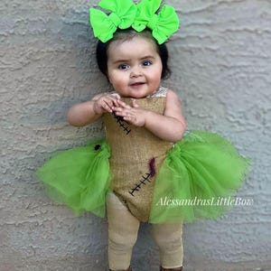 May include: A baby in a burlap-style costume with a green tulle skirt and a large green bow headband. The costume features stitched details and brown boots. The text "AlessandrasLittleBow" is visible.