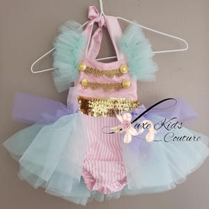 Könnte beinhalten: Ein Baby-Outfit im Zirkus-Stil mit einem rosa Oberteil, das mit goldenen Verzierungen und Knöpfen verziert ist. Das Outfit hat einen gestreiften Unterteil, einen goldfarbenen Pailletten-Gürtel und einen mehrschichtigen Tüllrock in Blau- und Lilatönen. Der Text "Luxe Kids Couture" ist sichtbar.