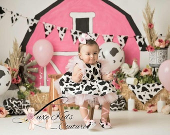 Könnte beinhalten: Ein Baby in einem schwarz-weiß Kuhmuster-Kleid mit rosa Schleife und Schuhen. Der Hintergrund zeigt eine rosa Scheune, Kuhmuster-Ballons und dekorative Elemente, die eine Bauernhof-Szene schaffen.