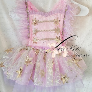 May include: A pink tulle tutu with gold star sequins and a pink bodice with gold trim and star embellishments. The bodice has a lavender feather trim on the straps.  "Luxe Kids Couture" is printed on the skirt.