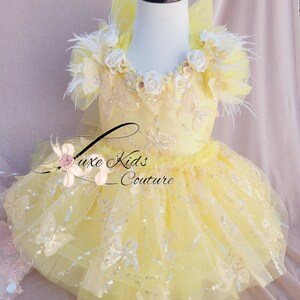 May include: A yellow tulle dress with a sequined bodice and butterfly embellishments. The dress has a white flower and feather trim around the neckline. The dress is on a white mannequin.