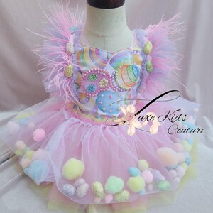 May include: A pink tulle tutu with pastel colored pom-poms and a white top with a colorful Easter egg design. The top has pink feathers on the shoulders.