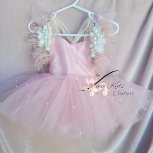 May include: A light pink tutu dress with a tulle skirt and pearl accents. The bodice features a V-neck design and is adorned with pearl and feather embellishments on the straps. The dress is hanging on a white hanger.