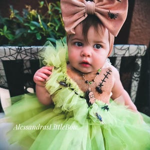 Könnte beinhalten: Ein Baby in einem Halloween-Kostüm. Das Outfit umfasst einen grünen Tüllrock, ein Oberteil aus Jute mit Insektenverzierungen und ein großes Schleifen-Stirnband mit Spinnendekorationen. Der Text "AlessandrasLittleBow" ist sichtbar.