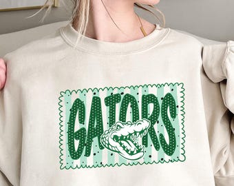 Op de afbeelding: Een beige sweater met ronde hals en een rechthoekige afbeelding met een geschulpte rand. Het ontwerp bevat lichtgroene verticale strepen, donkergroene stippen en het woord GATORS in grote letters met een illustratie van een alligatorkop.