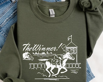 Könnte beinhalten: Ein olivgrünes Sweatshirt mit einer weißen Grafik einer Pferderennszene. Das Design zeigt ein Pferd, einen Jockey, eine Rennstrecke und den Text "The Winner!". Das Sweatshirt ist auf einer blauen Jeans gefaltet.