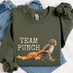 May include: An olive green sweatshirt with the text "TEAM PUNCH" and a graphic of two monkeys. One monkey is pulling the other by the arm. The sweatshirt is displayed on top of a pair of blue jeans.