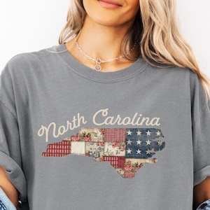Puede incluir: Camiseta gris con la inscripción "North Carolina" en letra cursiva. El contorno del estado está relleno con un diseño de patchwork de patrones florales, rojos y azules, que se asemeja a una bandera. La camiseta es de estilo casual.