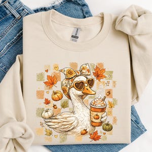 Pode incluir: Uma camisola bege de gola redonda com o gráfico de um pato branco a usar um lenço floral e óculos de sol em forma de coração. O pato segura um café com natas, rodeado por folhas de ácer cor de laranja, pequenas abóboras e um fundo axadrezado.