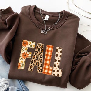 Peut inclure: Un sweat-shirt à col rond marron foncé avec le mot FALL en grandes lettres majuscules. Chaque lettre présente un motif unique : floral, léopard, carreaux orange et blanc, et pois marron. Un collier à chaîne argentée repose sur le col.