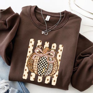 May include: A dark brown crewneck sweatshirt featuring a central graphic of a pumpkin with a light pink bow. The pumpkin has segments with leopard print, plaid, and checkered patterns, set against a background of vertical beige polka dot stripes.