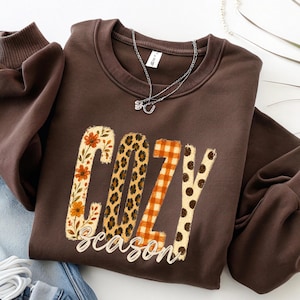 May include: A dark brown crewneck sweatshirt featuring the word COZY in large, textured letters with varied patterns including orange flowers, leopard print, orange plaid, and brown polka dots, with the word season written below.