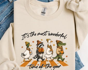 May include: A beige crewneck sweatshirt featuring a graphic of a duck, raccoon, goose, and alligator wearing autumn hats and scarves. The text reads, "It's the most wonderful time of the year," with orange leaves and a crosswalk design.
