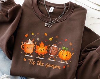 May include: A dark brown crewneck sweatshirt featuring a graphic print of a smiling orange mug, a maple leaf, a football, and a pumpkin with the text 'Tis the season' in cursive script. The sweatshirt has long sleeves with ribbed cuffs and a crew neckline.