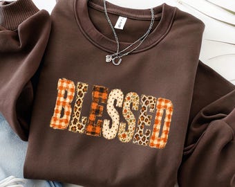 May include: A dark brown long-sleeve sweatshirt featuring the word BLESSED in large, block-style letters. Each letter has a unique pattern, including orange and white plaid, leopard print, and floral designs. A silver chain necklace rests on the collar.