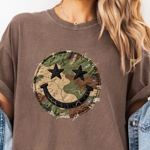 May include: Espresso-coloured t-shirt featuring a camouflage smiley face with black star eyes. The smiley face is a patch with a distressed, stitched edge. The shirt has a relaxed fit and rolled sleeves.