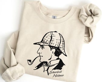 May include: A beige sweatshirt featuring a black illustration of Sherlock Holmes with a pipe and hat. The words "Sherlock Holmes" are printed below the illustration. The word "Natural" is written in cursive in the upper left corner.