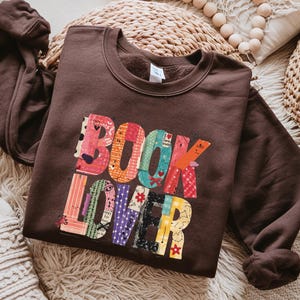 May include: A brown sweatshirt with the words "BOOK LOVER" in colorful, patchwork-style letters. The letters are made up of various patterns and textures, including floral, striped, and patterned designs. The sweatshirt has long sleeves and a crew neck.