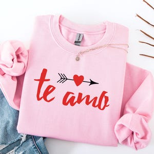 Op de afbeelding: Een lichtroze sweatshirt met de woorden "te amo" in rood schrift, met een hart doorboord door een pijl. Een zilveren ketting met een kleine hanger ligt op het sweatshirt. Het sweatshirt ligt op een spijkerbroek.