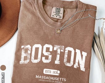 May include: Espresso-colored t-shirt with the word "BOSTON" in large white letters. Below, it reads "ESTD. 1630, MASSACHUSETTS, UNITED STATES OF AMERICA." A silver necklace with a pendant and a brown hat are also visible.