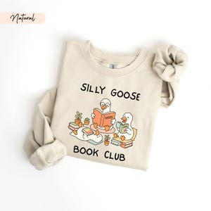 May include: A cream-coloured sweatshirt featuring the text "SILLY GOOSE BOOK CLUB" and a graphic of three cartoon geese reading books. One goose is reading a "NOVEL", another a "COMIC". The sweatshirt has long sleeves and a crew neck.