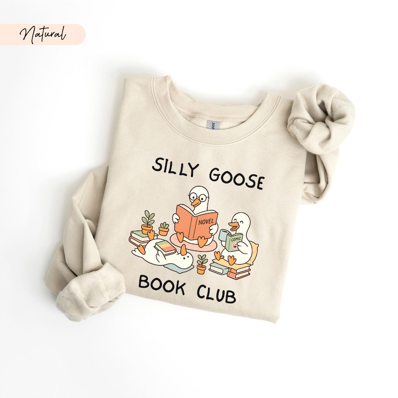 May include: A cream-coloured sweatshirt featuring the text "SILLY GOOSE BOOK CLUB" and a graphic of three cartoon geese reading books. One goose is reading a "NOVEL", another a "COMIC". The sweatshirt has long sleeves and a crew neck.