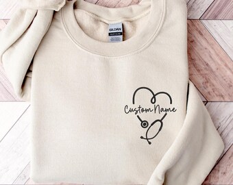 May include: A beige sweatshirt with a crew neck and long sleeves. A black heart-shaped design with a stethoscope is printed on the front, along with the words "Custom Name" in a script font. The sweatshirt is folded neatly.
