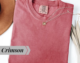 May include: A crimson short-sleeved t-shirt with a crew neckline and a Comfort Colors brand label inside the collar. A delicate silver-toned chain necklace with a small circular pendant featuring a rose design rests on the fabric.